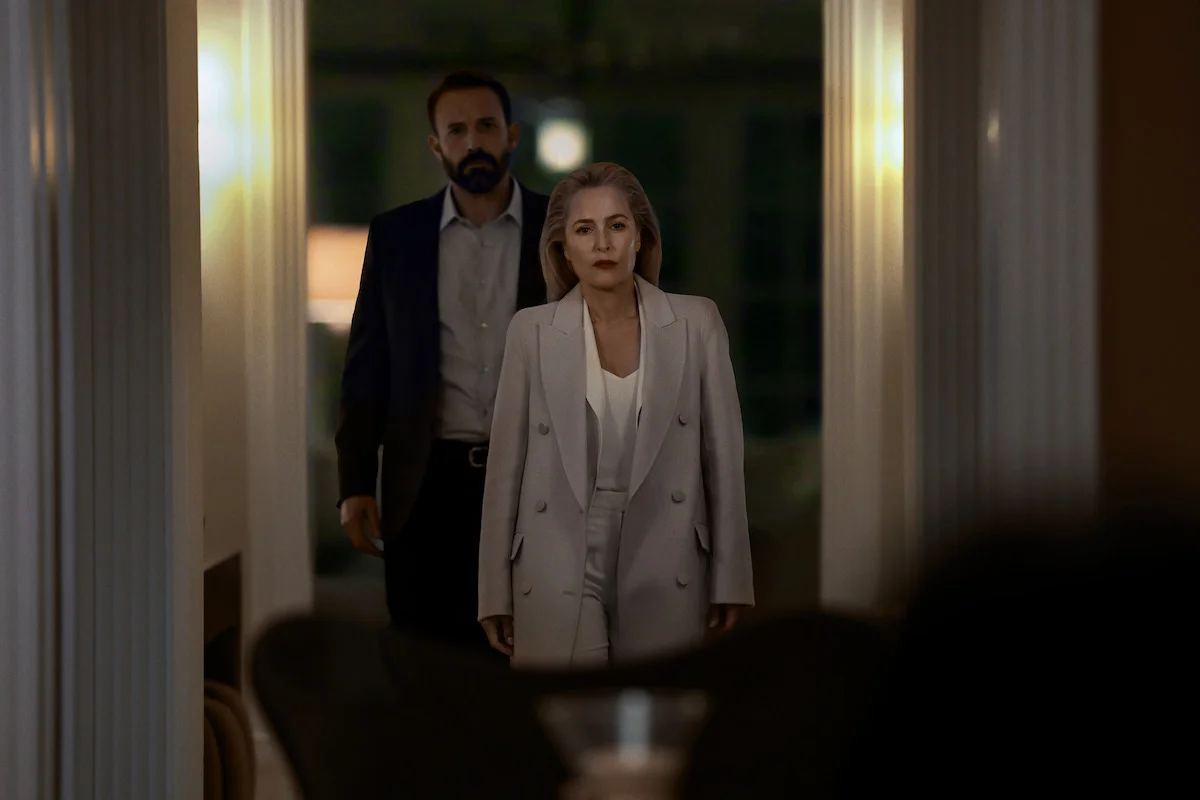 Gillian Anderson, con un traje claro, aparece en un pasillo con un hombre detrás.