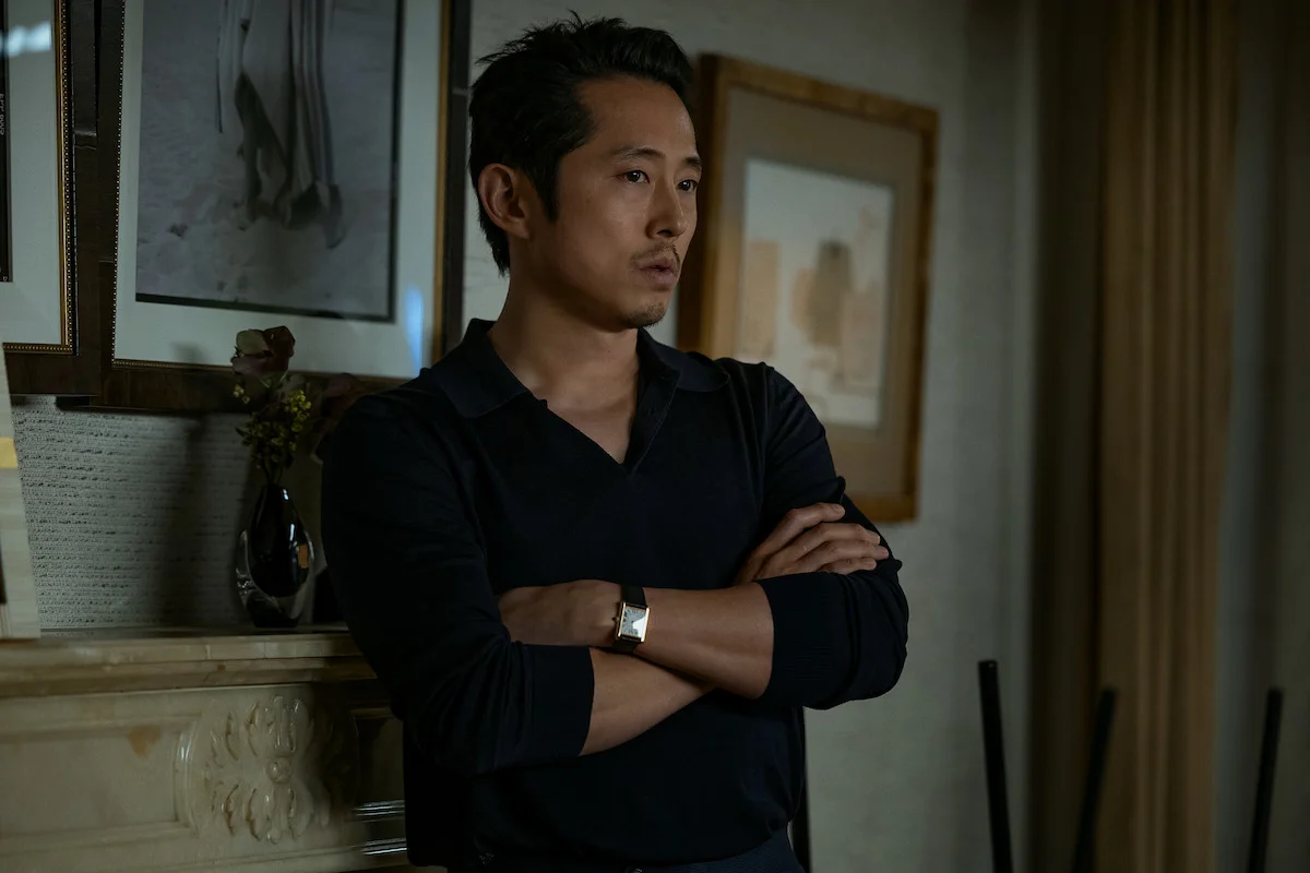 Steven Yeun está de pie con los brazos cruzados junto a una repisa con fotos enmarcadas.