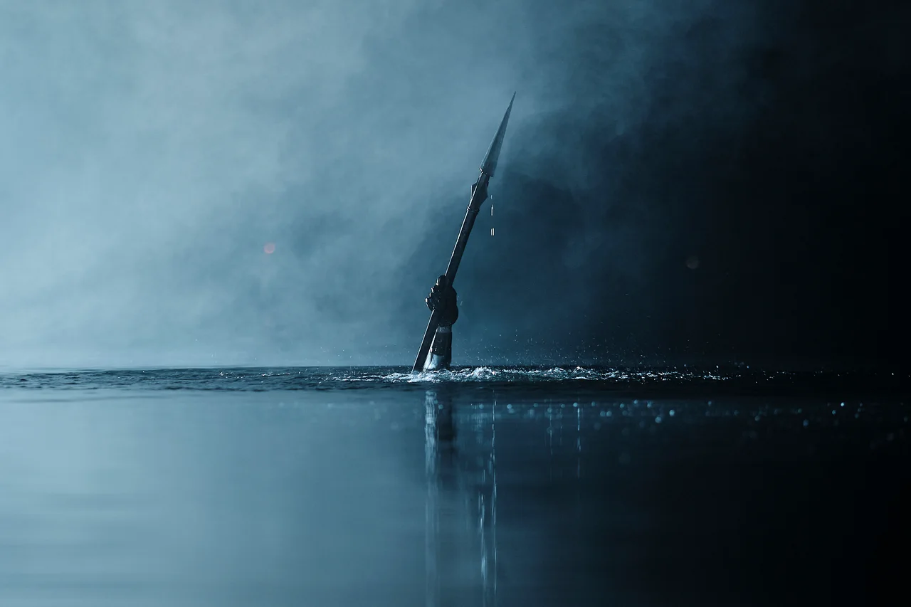Una figura sostiene un objeto largo en diagonal sobre el agua, entre niebla y árboles oscuros.
