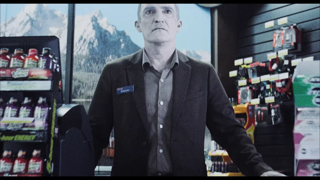 Un hombre con saco oscuro está en un comercio, entre exhibidores con paquetes de golosinas.