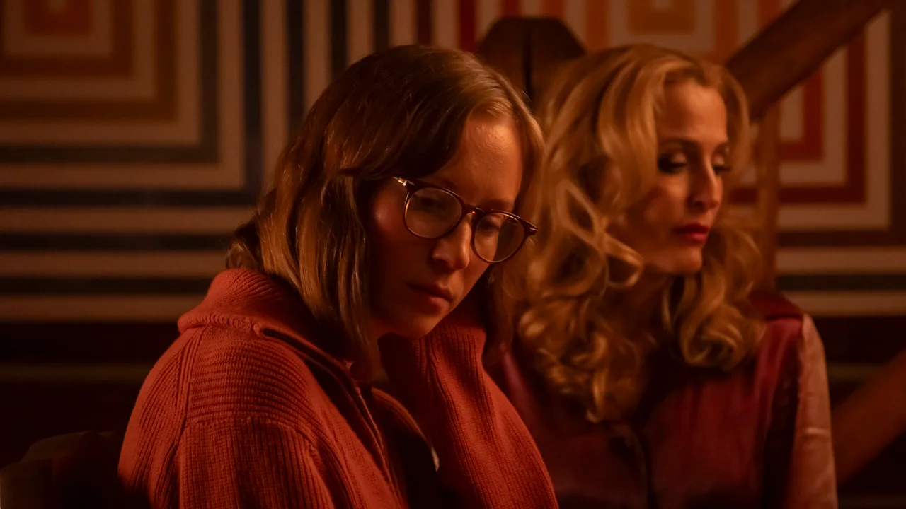 Hannah Einbinder, con anteojos y un sweater rojo, está junto a Gillian Anderson en un interior de madera.