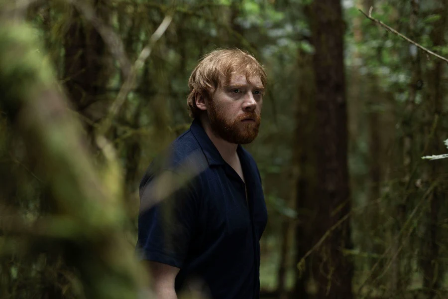 Rupert Grint mira hacia un costado entre los árboles del bosque.