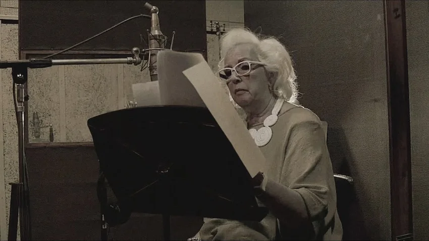 Susana Rinaldi, con anteojos blancos, lee una hoja frente a un micrófono en un estudio.
