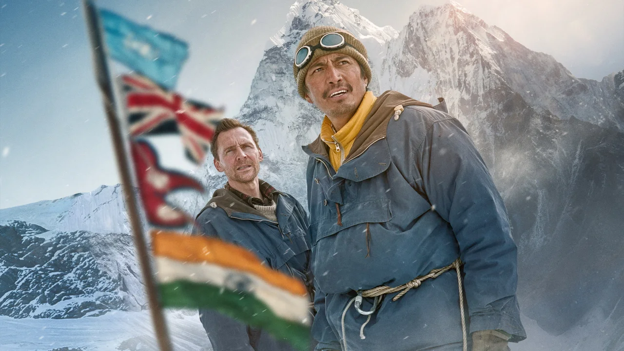 Dos montañistas con ropa de expedición miran hacia la montaña, junto a banderas, en Tenzing.