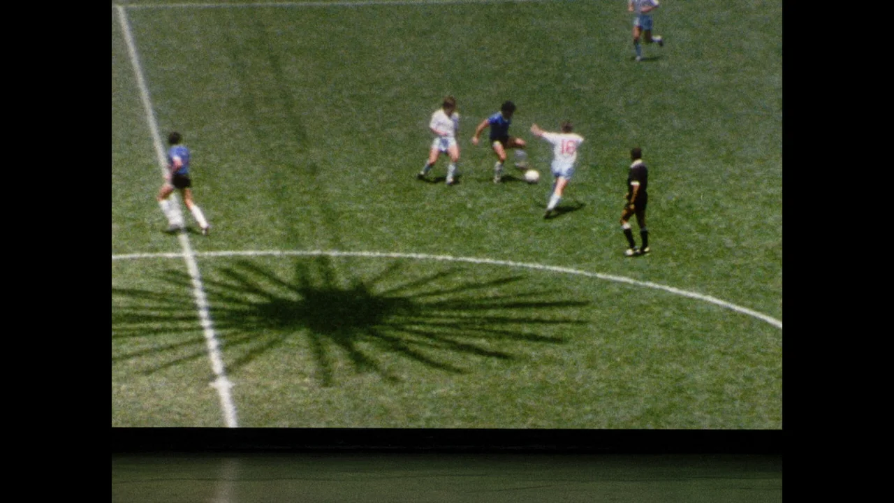 Un jugador de camiseta azul lleva la pelota por la cancha mientras un rival con camiseta blanca intenta marcarlo.