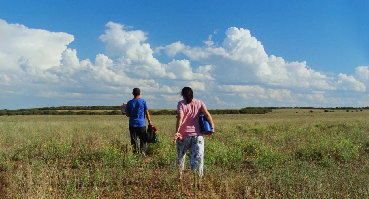 Dos personas caminan de espaldas por un campo abierto bajo un cielo con nubes.