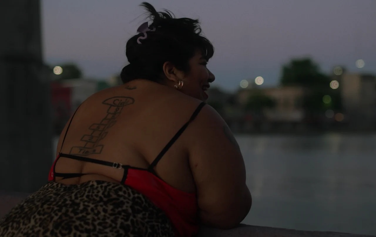 Una mujer de espaldas gira el rostro y sonríe, con edificios desenfocados al atardecer.