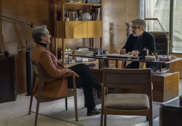 Diego Peretti, con saco color salmón, conversa con Ricardo Darín a ambos lados de un escritorio.