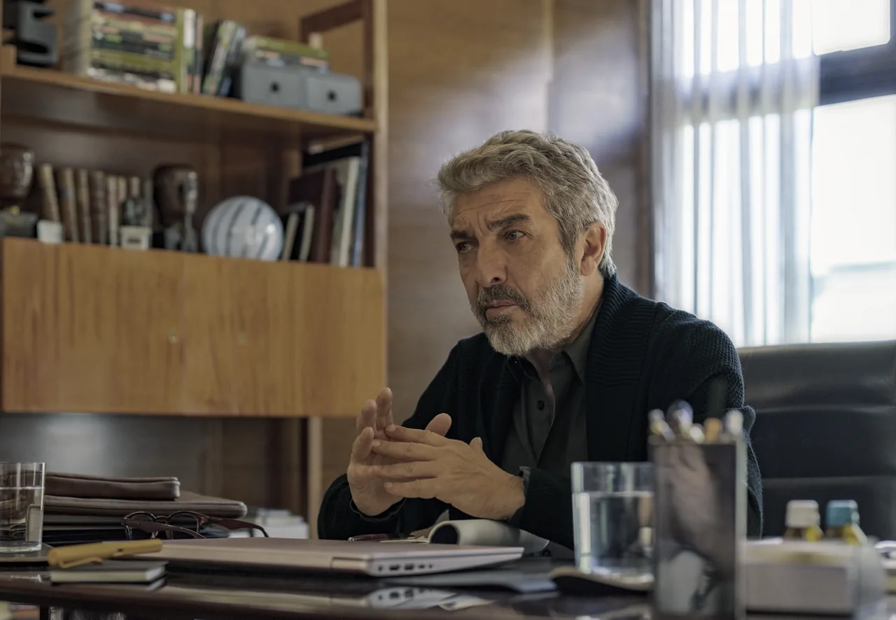 Ricardo Darín está sentado detrás de un escritorio, con las manos juntas y una ventana a su derecha.