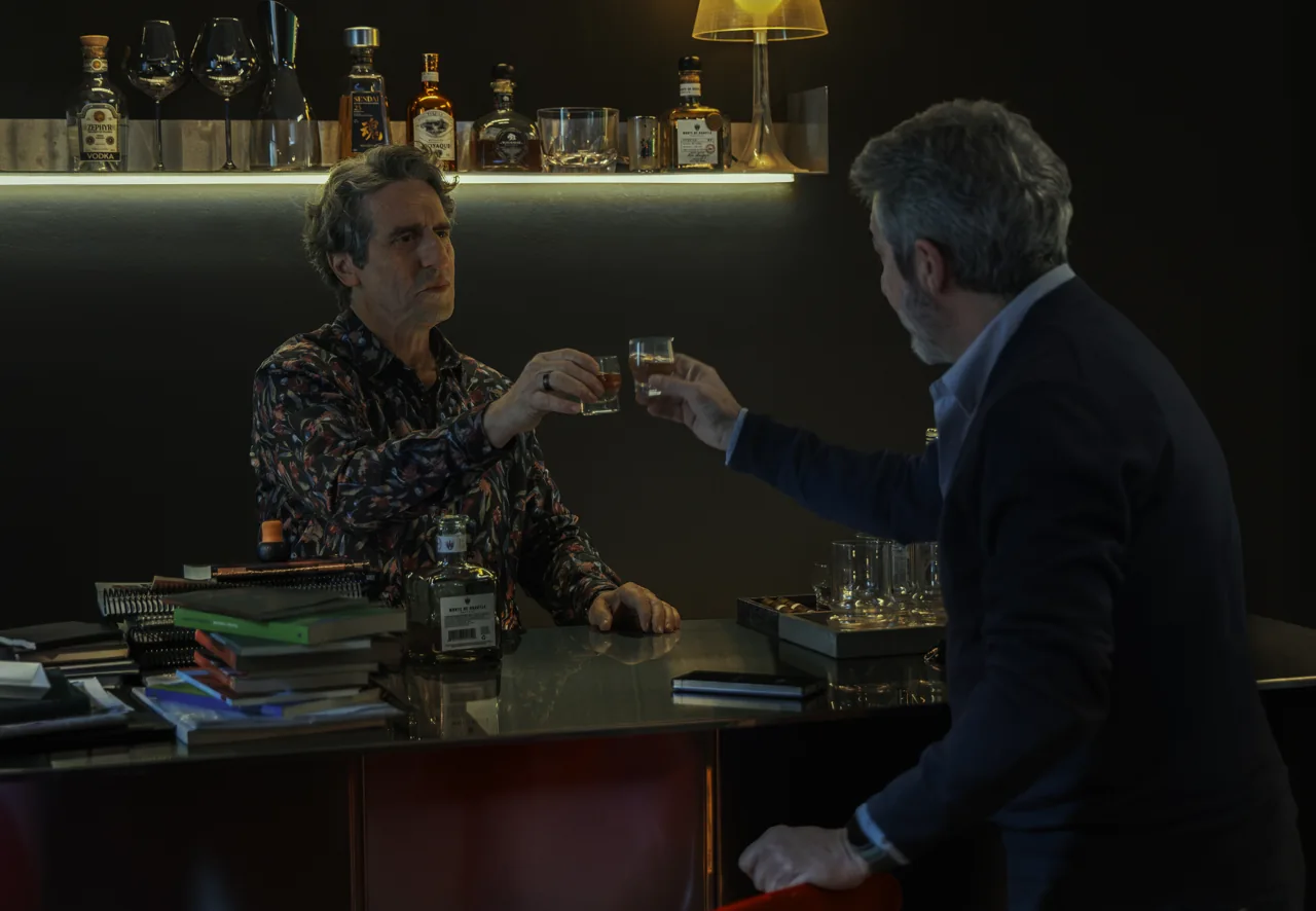 Diego Peretti y Ricardo Darín acercan sus vasos en una barra rodeada de botellas y libros.