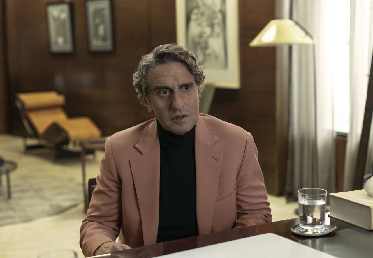 Diego Peretti lleva un saco color salmón y un suéter negro, sentado junto a un vaso de agua.
