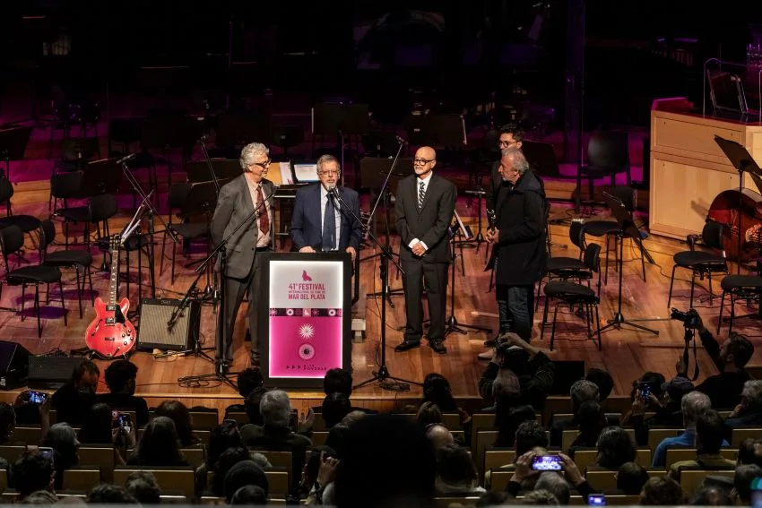 Cuatro hombres están de pie en un escenario con atril e instrumentos, frente al público sentado.