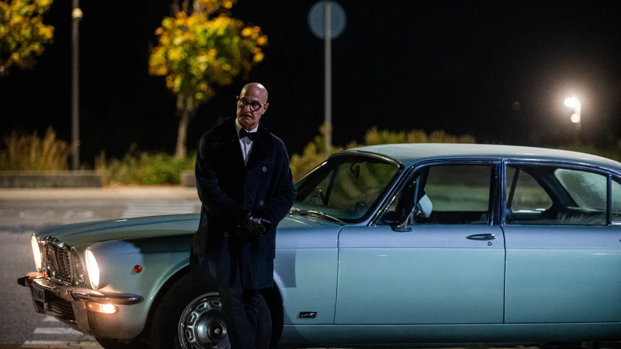 Stanley Tucci, vestido de traje oscuro, está junto a un auto celeste de noche.