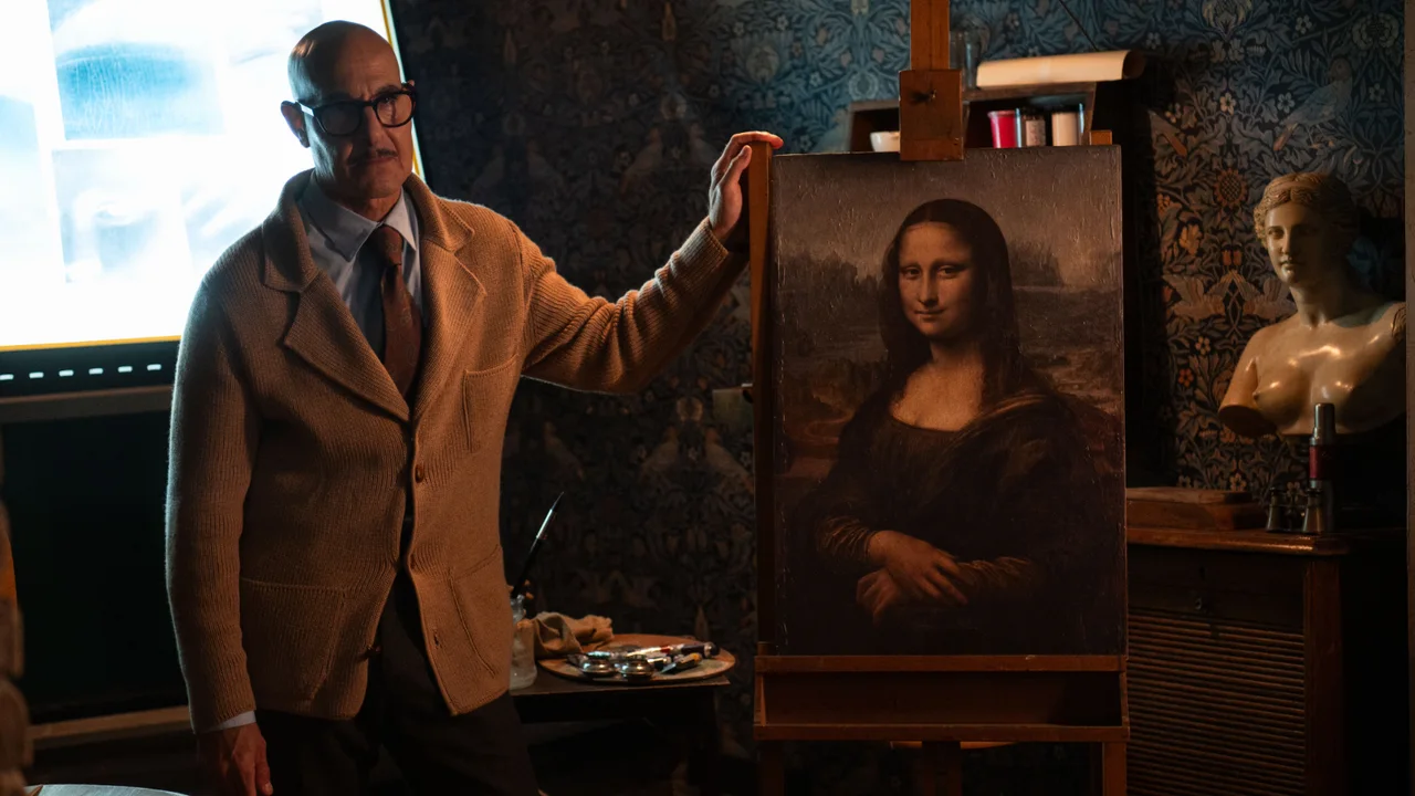 Stanley Tucci, con anteojos y saco claro, apoya una mano en el marco de una reproducción de la Mona Lisa.