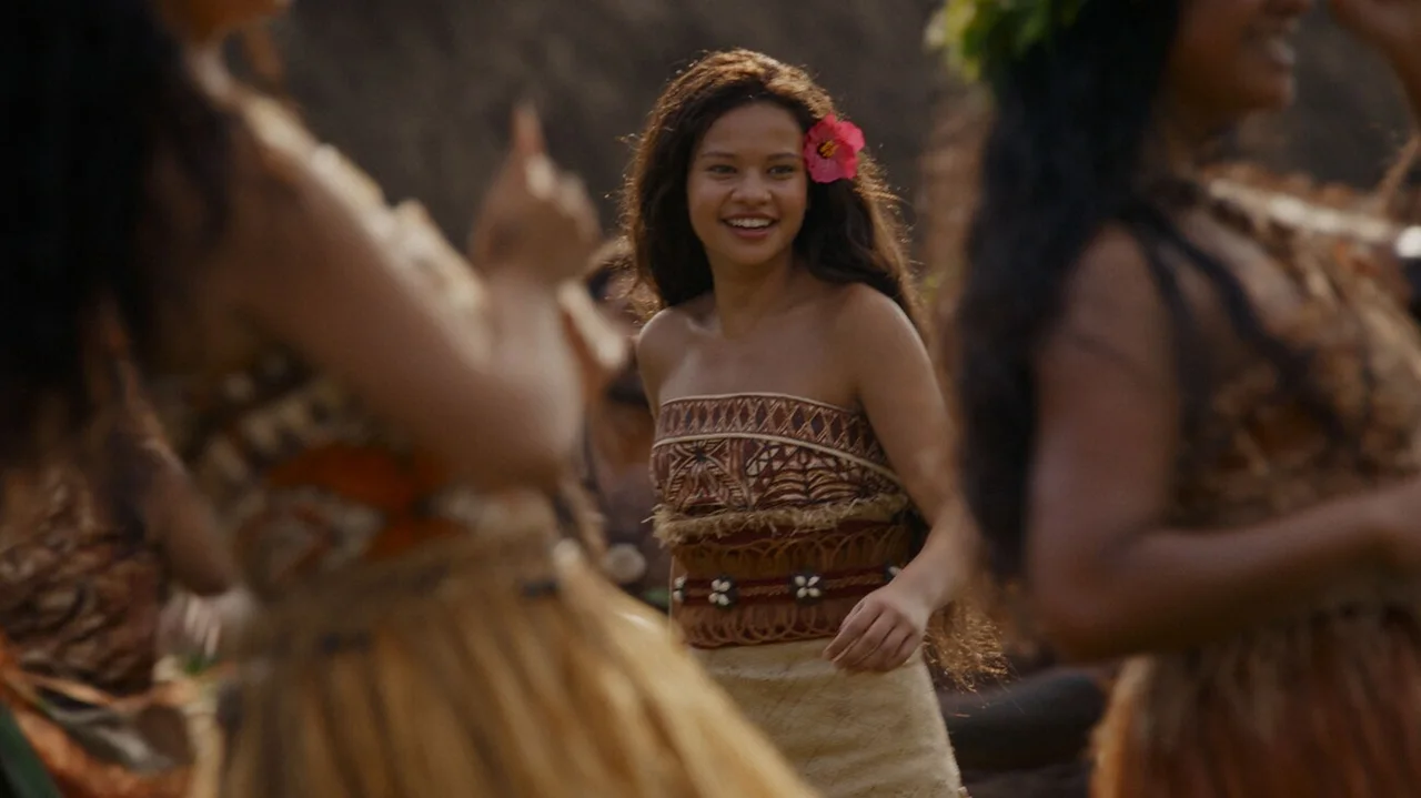 Moana sonríe con una flor en el pelo, rodeada de mujeres que llevan collares y ropa de la isla.