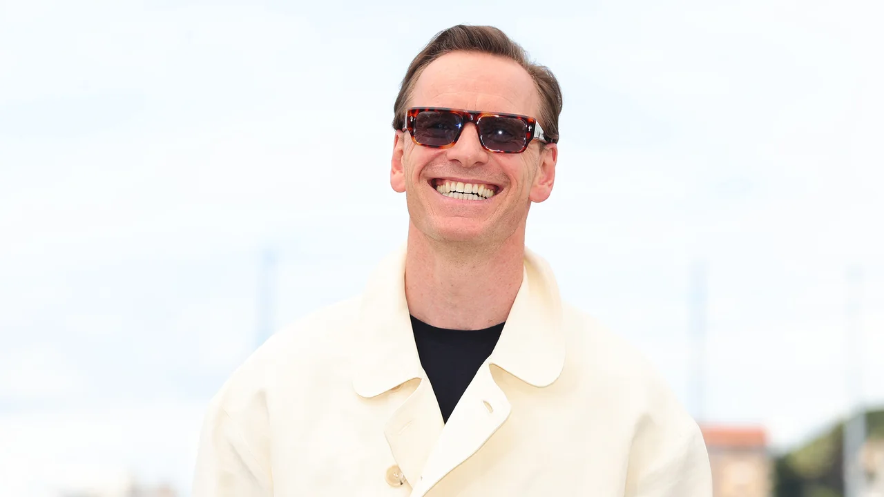 Michael Fassbender sonríe con anteojos de sol y un saco claro frente al fondo del photocall.