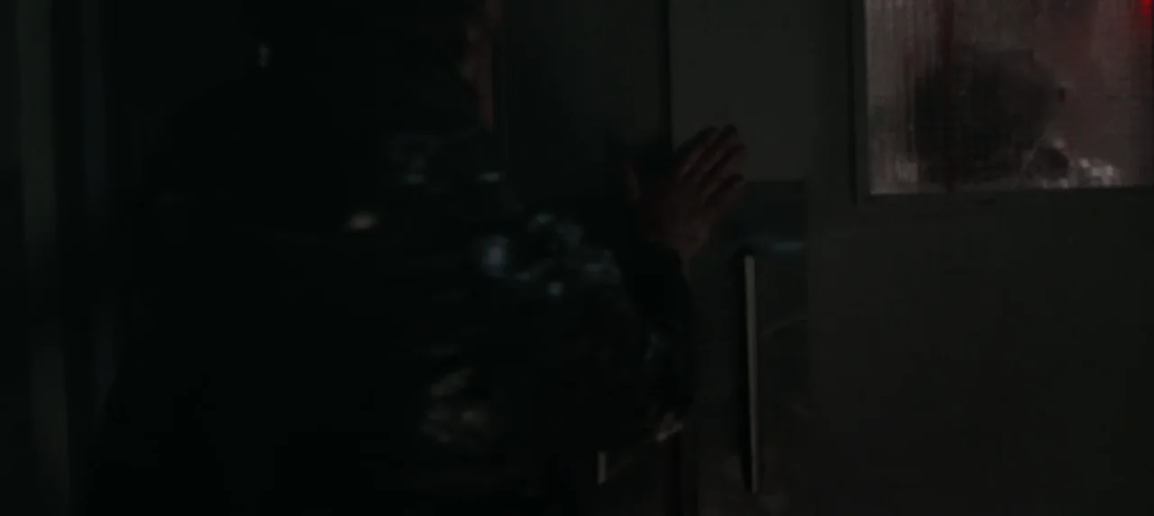 Bryan apoya las manos contra una puerta con una ventana pequeña y luz roja al otro lado.
