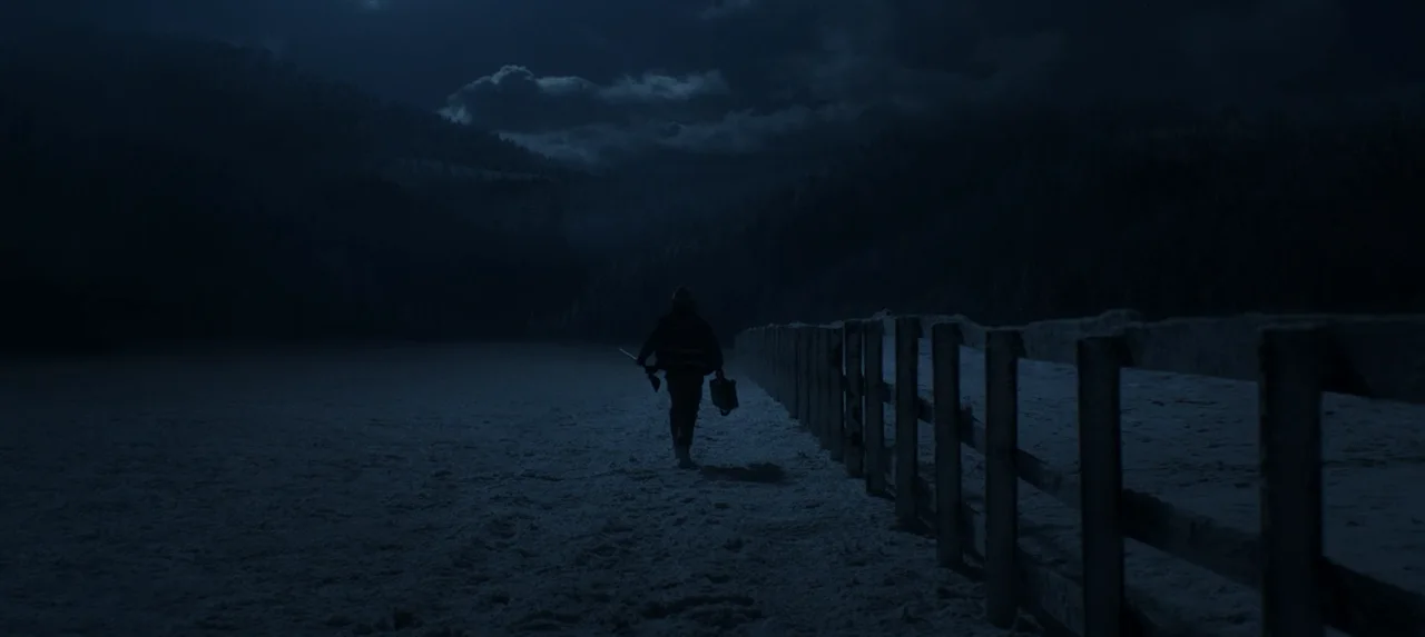 Una figura avanza de noche junto a una baranda, bajo un cielo cubierto de nubes.