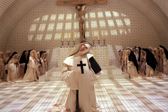 Una religiosa está en el centro de un salón blanco abovedado, rodeada por otras monjas.