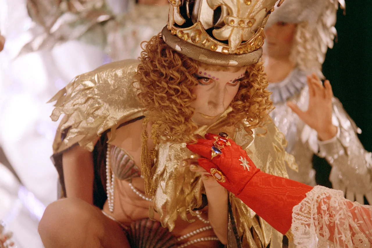 Un personaje con una corona y vestuario dorado sostiene un objeto pequeño junto a una figura con un guante rojo.